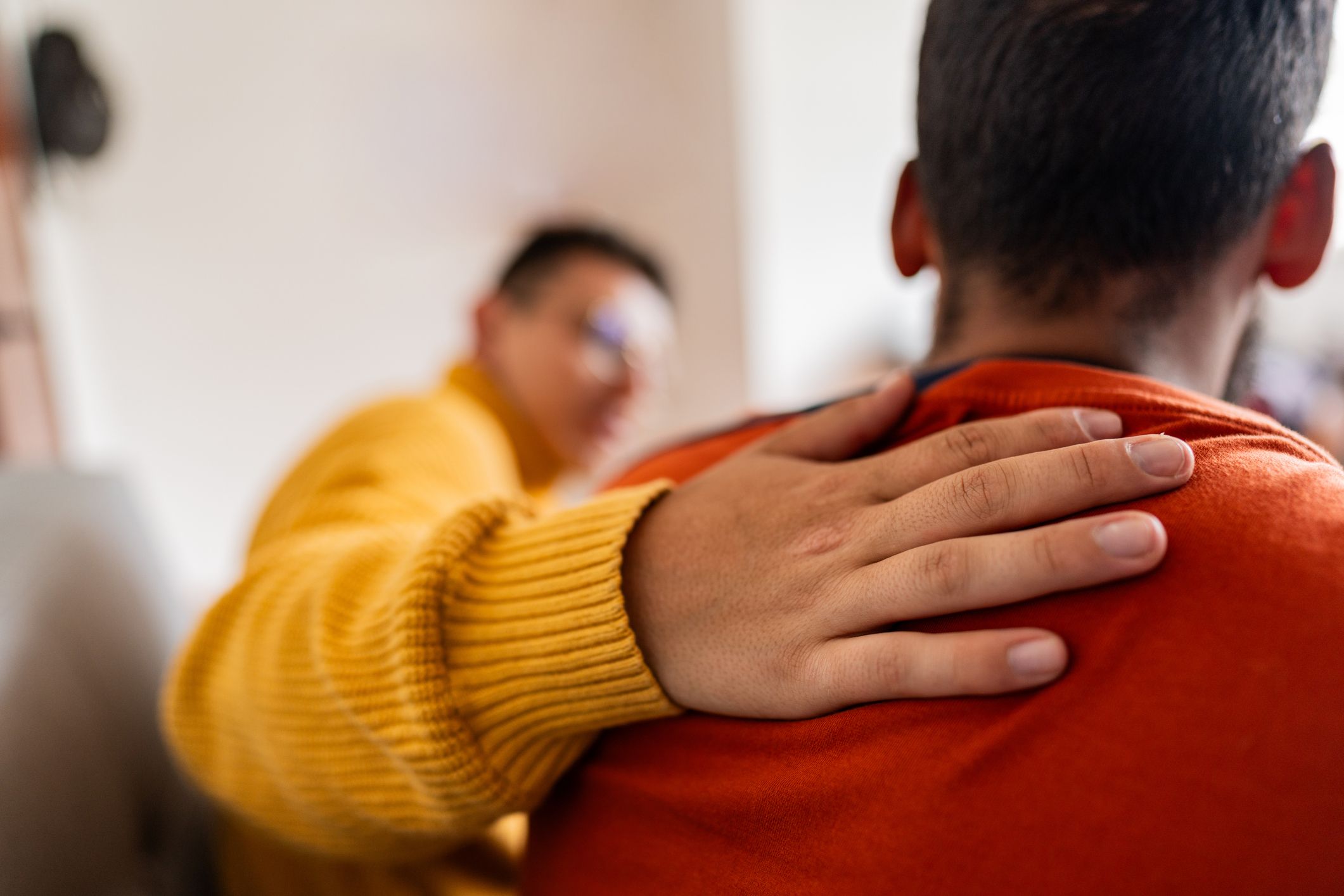 Eine Hand in einem gelben Ärmel liegt tröstend auf der Schulter einer Person im orangefarbenen Shirt. Im Hintergrund ist unscharf eine weitere Person mit Brille zu erkennen.